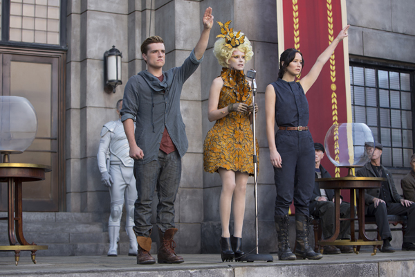 Peeta (Hutcherson) and Katniss (Lawrence) give the rebellion 3 finger salute after being introduced by Effie Trinket (Banks)